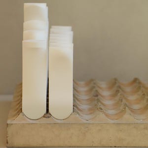 Op de afbeelding: Een verzameling witte, rechthoekige objecten met gebogen toppen, mogelijk kaarsen of zeep, gerangschikt op een beige basis met golvende randen. De objecten zijn in twee groepen gestapeld, wat een minimalistische, strakke esthetiek cre&euml;ert.
