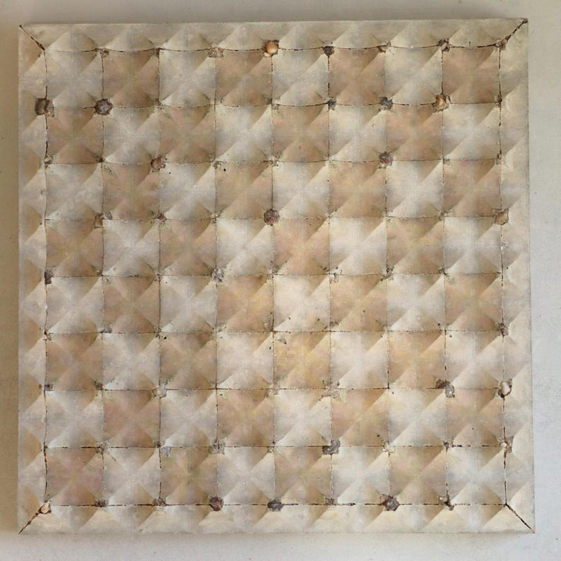 Op de afbeelding: Vierkant kunstwerk met een herhalend patroon van verhoogde, ruitvormige secties. Het oppervlak heeft een getextureerde, verouderde uitstraling met beige, bruin en bruin tinten. Kleine, donkere vlekken zijn verspreid over het oppervlak.