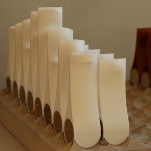 Op de afbeelding: Een verzameling witte, abstracte architectuurmodellen. De modellen zijn in een rij gerangschikt, met verschillende hoogtes en vormen. Elk model heeft een afgeronde bovenkant en een smalle, langwerpige basis. De bases zijn van een bruin, kurkachtig materiaal.