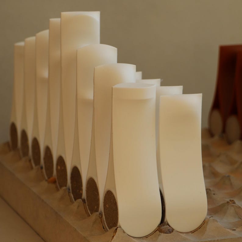 Op de afbeelding: Een verzameling witte, abstracte architectuurmodellen. De modellen zijn in een rij gerangschikt, met verschillende hoogtes en vormen. Elk model heeft een afgeronde bovenkant en een smalle, langwerpige basis. De bases zijn van een bruin, kurkachtig materiaal.