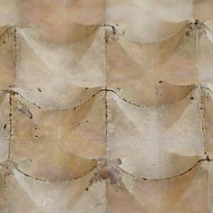 Op de afbeelding: Close-up van een getextureerd, lichtbruin oppervlak met een herhalend patroon van gebogen en hoekige vormen. Het oppervlak lijkt te zijn gemaakt van een ruw materiaal, mogelijk beton of steen, met zichtbare onvolkomenheden.