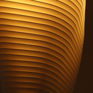 May include: Abstract image of a cylindrical object with horizontal, curved lines. The object has a warm, golden-yellow glow, with darker shadows creating depth. The design suggests a modern, architectural theme.