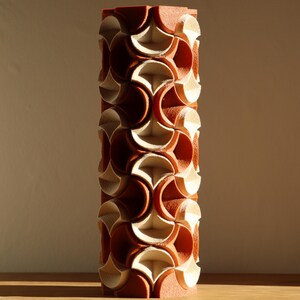 May include: A tall, cylindrical decorative object with a repeating pattern of interlocking shapes. The object is primarily reddish-brown with cream-colored accents, creating a textured, geometric design. The piece is standing on a wooden surface.