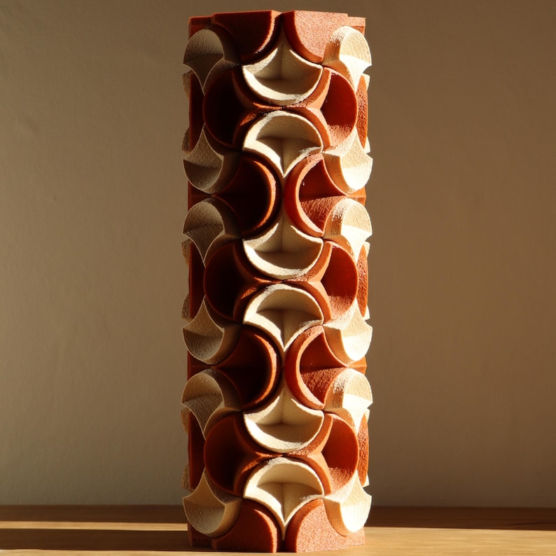 May include: A tall, cylindrical decorative object with a repeating pattern of interlocking shapes. The object is primarily reddish-brown with cream-colored accents, creating a textured, geometric design. The piece is standing on a wooden surface.