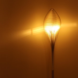 May include: A tall, modern floor lamp with a unique design. The lamp features a spiraling, layered shade that emits a warm, golden light. The base is a slender, metallic pole. The background is a solid, warm-toned wall.