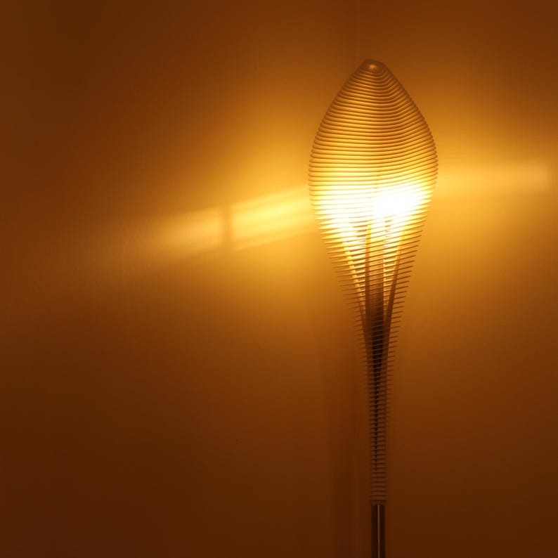 May include: A tall, modern floor lamp with a unique design. The lamp features a spiraling, layered shade that emits a warm, golden light. The base is a slender, metallic pole. The background is a solid, warm-toned wall.