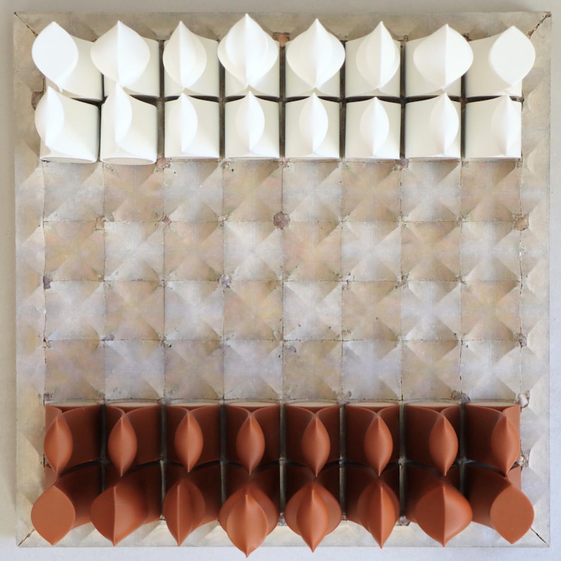 Op de afbeelding: Abstracte wandkunst met een geometrisch ontwerp. Het kunstwerk toont een raster van vierkante tegels in een neutrale kleur, met witte en terracotta-kleurige driedimensionale vormen gerangschikt in rijen. Het stuk heeft een getextureerd uiterlijk en is gemonteerd op een lichtgekleurd oppervlak.
