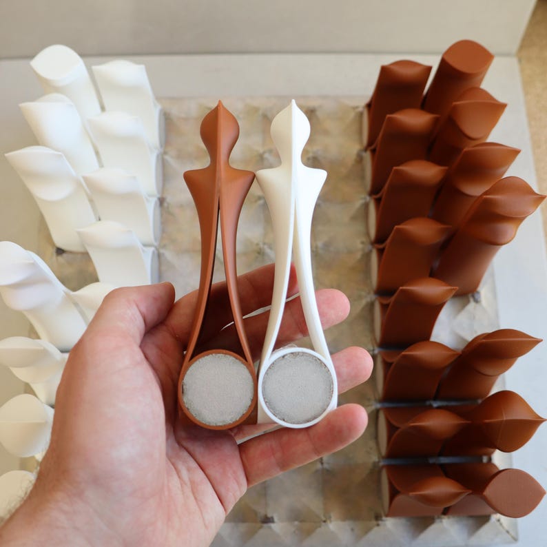Op de afbeelding: Een modern schaakspel met witte en bruine stukken. Twee stukken worden in een hand gehouden, &eacute;&eacute;n wit en &eacute;&eacute;n bruin. De stukken hebben een uniek, gestileerd ontwerp en zijn gerangschikt op een raster.