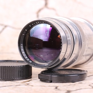 Può includere: Un obiettivo per fotocamera argentato con segni neri. L'obiettivo ha un tappo nero e un tappo dell'obiettivo nero. L'obiettivo è su una superficie testurizzata marrone chiaro.