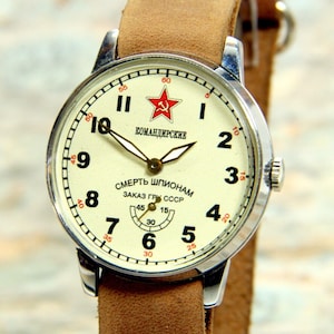 May include: A vintage silver wristwatch with a white face and black numbers. The watch has a red star and the text "КОМАНДИРСКИЕ" and "СМЕРТЬ ШПИОНАМ ЗАКАЗ ГР СССР" on the face.