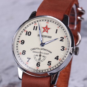 Puede incluir: Un reloj de pulsera plateado con una esfera blanca y agujas negras. La esfera del reloj tiene números rojos y una estrella roja con un martillo y una hoz. La esfera del reloj también tiene el texto "СМЕРТЬ ШПИОНАМ ЗАКАЗ ПРУ СССР" en escritura cirílica.