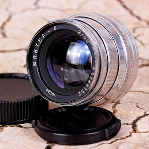 May include: A silver and black camera lens with the text "ЮПИТЕР" and "2/50" printed on it. The lens has a series of numbers printed around the edge, including "M 1 2 4 5.6 8 11 16 22 1.5 17".