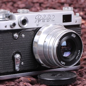 Puede incluir: Una cámara de película de 35 mm de color plateado y negro con una lente negra. La cámara tiene una cubierta de imitación de cuero negro y un cuerpo de metal plateado. La cámara tiene una tapa de lente negra.