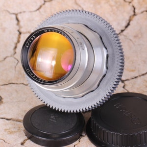 May include: A silver camera lens with a black ring and a gear-like ring around the outside. The lens has the text "РОНТЕПТ-1" and "4/135" printed on it. The lens is sitting on a brown surface.