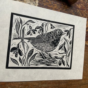 Puede incluir: Grabado en lin&oacute;leo en blanco y negro de un petirrojo posado en una rama con campanillas de invierno. El p&aacute;jaro est&aacute; mirando hacia la derecha y tiene un dise&ntilde;o estilizado y detallado.