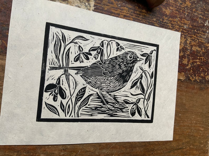 Puede incluir: Grabado en lin&oacute;leo en blanco y negro de un petirrojo posado en una rama con campanillas de invierno. El p&aacute;jaro est&aacute; mirando hacia la derecha y tiene un dise&ntilde;o estilizado y detallado.