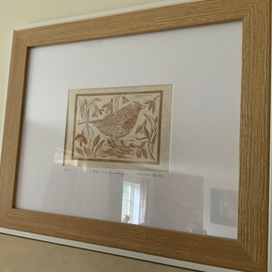 Puede incluir: Una impresi&oacute;n enmarcada de una linograf&iacute;a marr&oacute;n que representa un petirrojo posado en una rama con campanillas de invierno. La impresi&oacute;n est&aacute; titulada "Robin and Snowdrops" y firmada por "Miriam Cook".