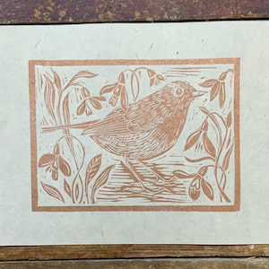 Puede incluir: Una impresi&oacute;n en lin&oacute;leo marr&oacute;n de un p&aacute;jaro posado en una rama con flores. La impresi&oacute;n est&aacute; en papel color crema.