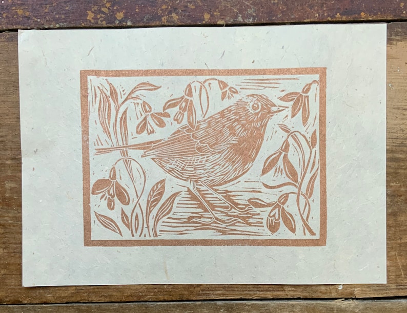 Puede incluir: Una impresi&oacute;n en lin&oacute;leo marr&oacute;n de un p&aacute;jaro posado en una rama con flores. La impresi&oacute;n est&aacute; en papel color crema.