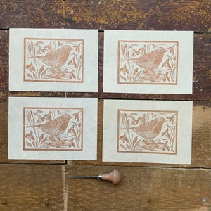Puede incluir: Cuatro tarjetas de papel marr&oacute;n con una impresi&oacute;n de lin&oacute;leo marr&oacute;n de un p&aacute;jaro en un campo de flores. Una peque&ntilde;a herramienta de talla de madera est&aacute; en primer plano.