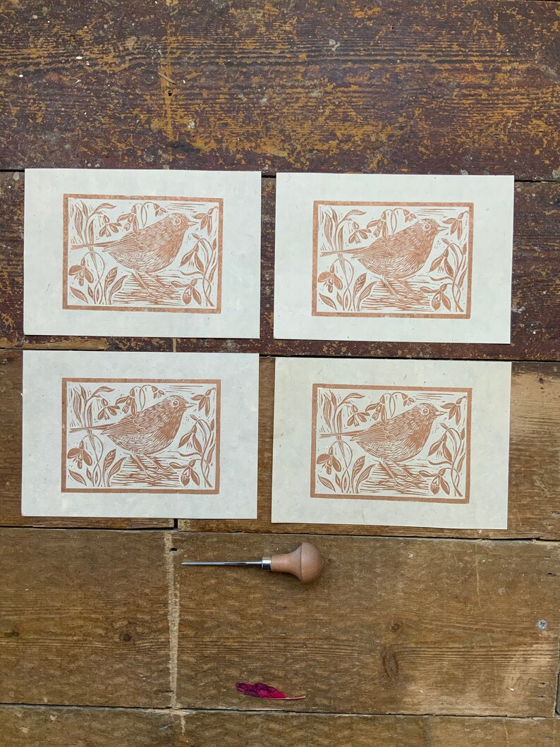 Puede incluir: Cuatro tarjetas de papel marr&oacute;n con una impresi&oacute;n de lin&oacute;leo marr&oacute;n de un p&aacute;jaro en un campo de flores. Una peque&ntilde;a herramienta de talla de madera est&aacute; en primer plano.