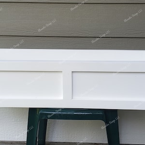 May include: White wooden window box planter with two divided compartments. The planter is sitting on a green stool.