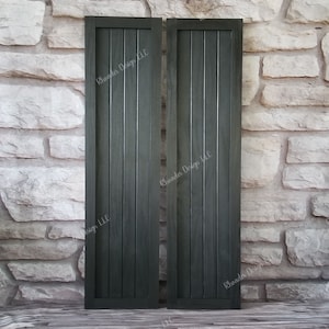May include: Two dark gray wooden shutters with vertical slats. The shutters are closed and have a rustic look. The text "Rhoades Design LLC" is printed on each shutter.