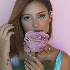 Puede incluir: Una mujer con cabello casta&ntilde;o sostiene una rosa rosa frente a su rostro. Lleva un top azul claro sin mangas y collares de oro. El fondo es rosa claro.
