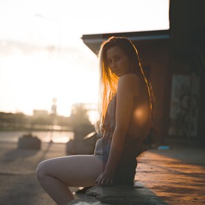 Puede incluir: Una mujer con el pelo largo y rubio se sienta en un borde de concreto, vestida con un vestido azul claro sin mangas y mirando por encima del hombro a la c&aacute;mara. El sol se pone detr&aacute;s de ella, proyectando un brillo c&aacute;lido en la escena.
