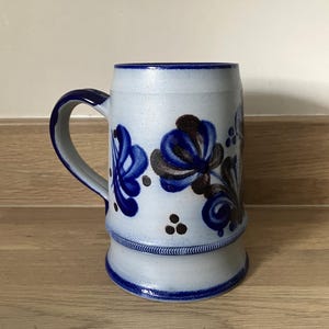 May include: A white ceramic beer stein with a blue and brown floral design. The stein has a handle and a wide base.