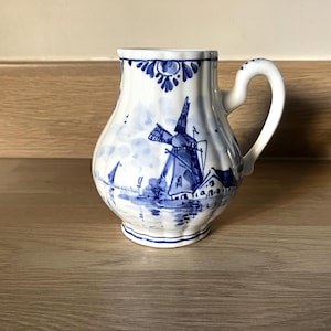 May include: A white and blue ceramic jug with a windmill scene. The jug has a curved handle and a rounded body. The windmill design is painted in blue on a white background. This jug is likely for decorative purposes.