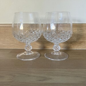 May include: Two clear crystal brandy glasses with diamond-cut patterns on the bowl. The glasses have a rounded bowl shape, a short stem, and a wide base. They are displayed on a wooden surface.