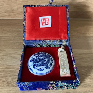 May include: A blue and white ceramic dragon-patterned circular seal paste container and a rectangular stone seal with carved characters, presented in a blue fabric-covered box with a red velvet interior and a white square label with red text.