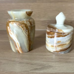 May include: Two decorative onyx stone pieces. One is a small vase with a flared top, and the other is a small jar with a lid. The pieces have a natural pattern of white, tan, and brown swirls.