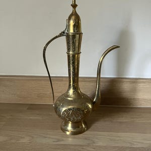 May include: A vintage brass pitcher with an ornate design. The pitcher has a long, curved spout, a handle, and a decorative lid. The body of the pitcher is engraved with floral patterns. The pitcher is standing on a wooden surface.