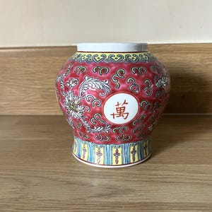 May include: A round, red ceramic jar with a white rim and base. The jar is decorated with white floral patterns and a circular emblem with a Chinese character. The base has a blue and yellow design.