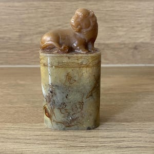 May include: A carved stone seal with a reclining tiger figurine on a cylindrical base. The base is decorated with etched imagery and Chinese characters. The seal is a light brown colour.