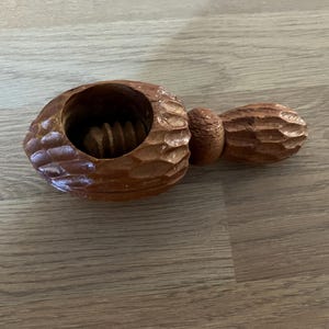 May include: A brown wooden nutcracker shaped like an acorn with a textured, carved exterior. The device features a central circular opening with a threaded screw mechanism inside, designed to crack nuts by twisting the handle attached to the side.