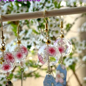 以下が含まれることがあります： 透明なガラス球のチャームにピンクの桜の花が咲き、青い花柄のチャームがぶら下がっているイヤリングのペア。イヤリングには「桜」と「Wind Chime」のラベルが貼られています。