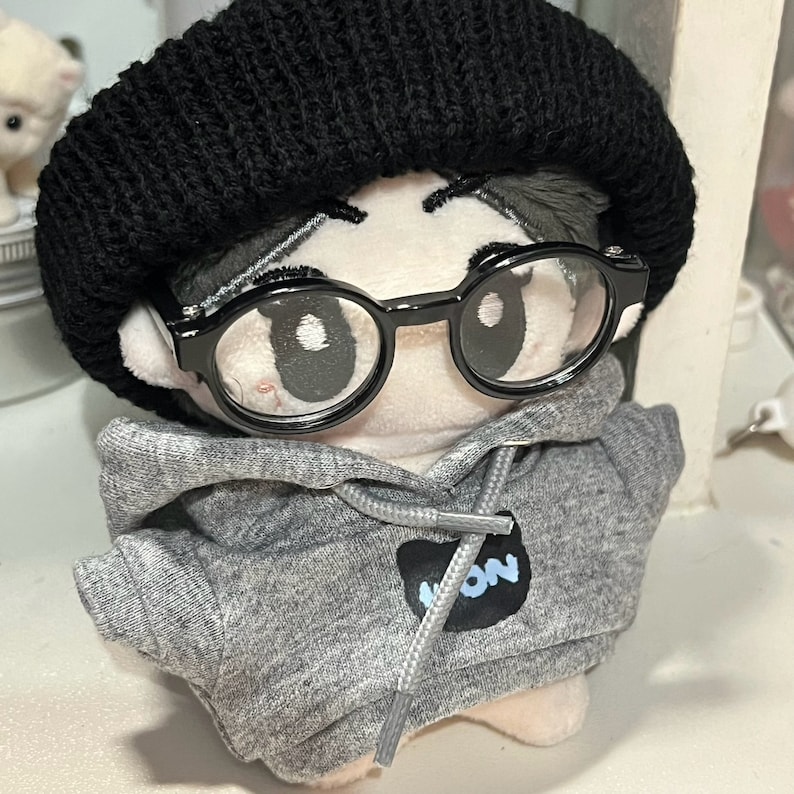 May include: A plush toy wearing a black beanie, grey hoodie with a black patch that says "NON", and black framed glasses.