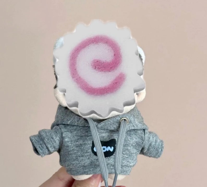 May include: A plush toy wearing a gray hoodie with a white logo that says "ION". The toy has a pink and white swirl pattern on its face.