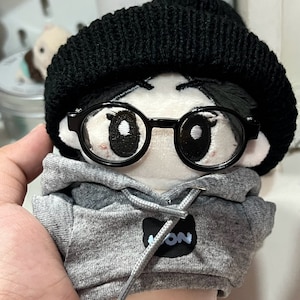 May include: A plush toy with a gray hoodie, black beanie, and black framed glasses. The hoodie has a black patch with the word "ION" on it.