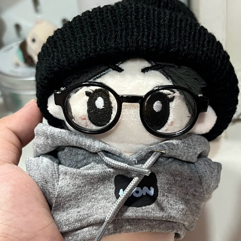 May include: A plush toy with a gray hoodie, black beanie, and black framed glasses. The hoodie has a black patch with the word "ION" on it.