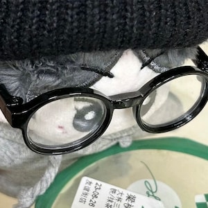 May include: A gray and white plush toy wearing a black beret and round black glasses. The toy has a stitched-on frown and a tag that reads "23-06-26  某某商店".