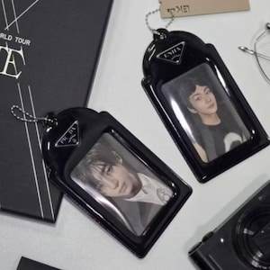 Puede incluir: Dos porta-credenciales negros con cadenas plateadas y ventanas transparentes, cada uno con una fotografía. Los porta-credenciales tienen un logotipo triangular y la palabra "ENHA" impresa. Una caja negra con texto blanco está en el fondo.