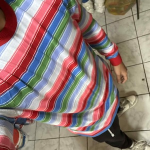 Könnte beinhalten: Ein langärmliges Hemd mit einem Streifenmuster in Rot, Weiß, Blau, Grün und Rosa. Das Hemd hat einen roten Rundhalsausschnitt und Manschetten. Die Person trägt eine schwarze Hose und weiße Turnschuhe.
