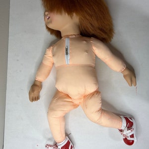 Op de afbeelding: Een pluche pop met rood haar, die een perzikkleurige jumpsuit en rode en witte sneakers draagt. De pop heeft een rits aan de voorkant van de jumpsuit.