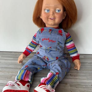 May include: A red-haired doll with blue eyes and a mischievous grin. The doll is wearing blue overalls with a red and white striped shirt and red and white sneakers. The overalls have a print of a fire hydrant and the words "Good Guys" on the front.
