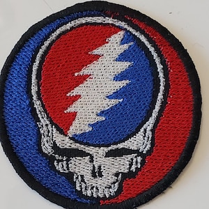 May include: Embroidered patch featuring the Steal Your Face skull logo. The patch has a circular shape with a black border. The design includes a red and blue color scheme with a white skull and lightning bolt.