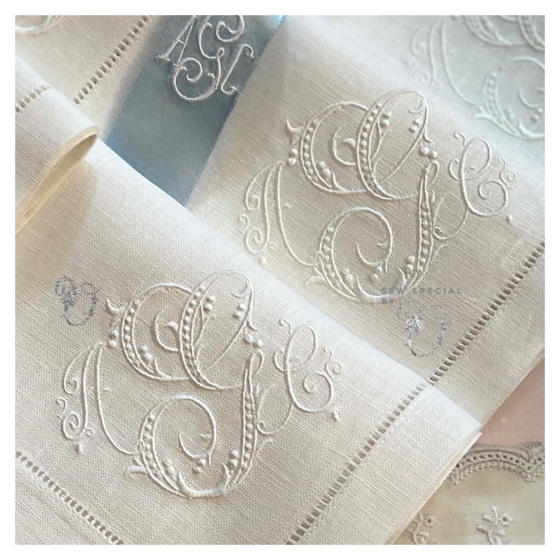 May include: Ivory linen napkins with embroidered initials. The napkins feature ornate, cursive monogram designs in a matching ivory thread. A light blue ribbon with embroidered initials is also visible.